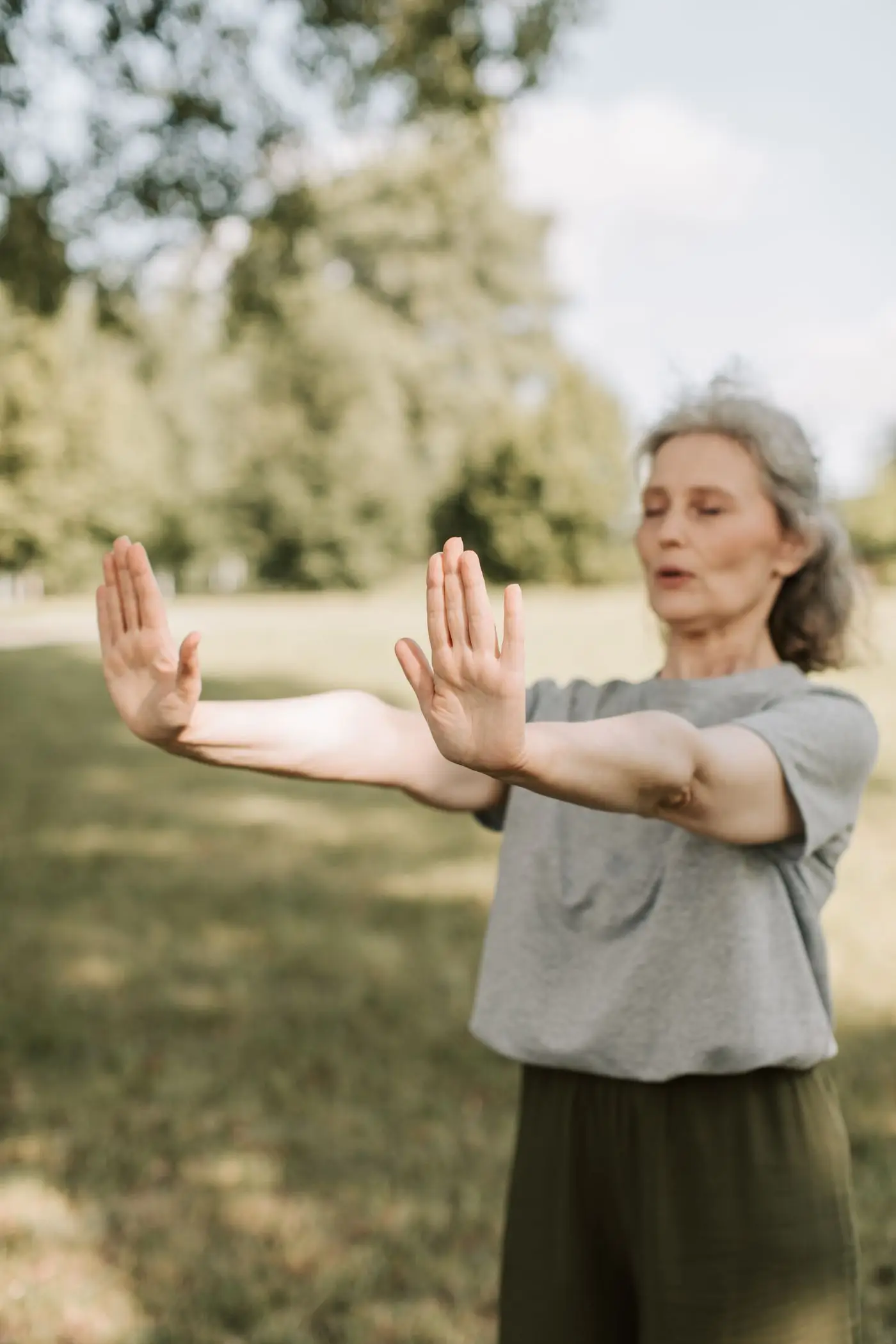 Une femme âgée dans un parc verdoyant, bras tendus et yeux fermés, tenant une pose d'équilibre paisible.